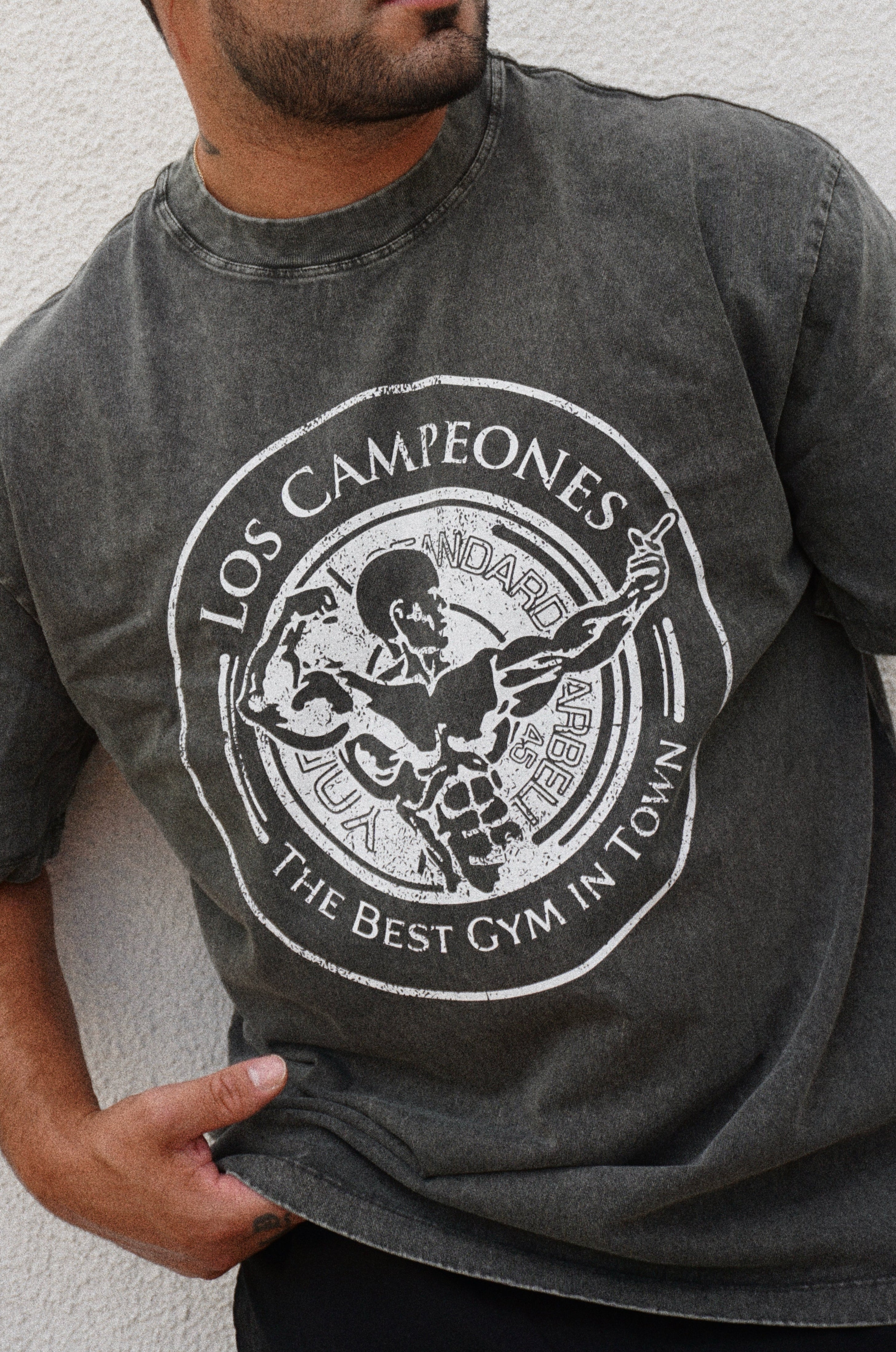 Man wearing a gray t-shirt with 'Los Campeones' graphic design against a white wall.